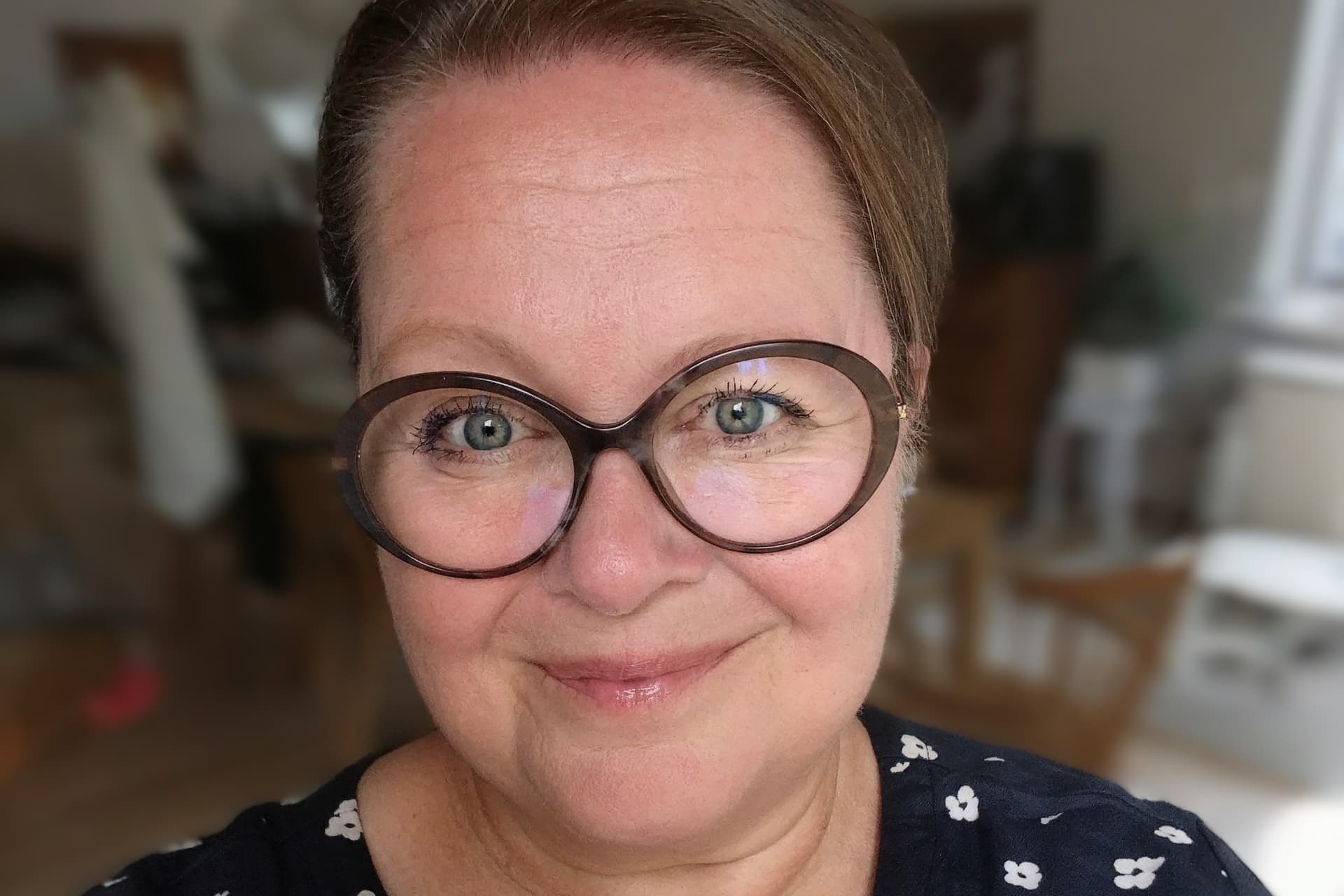 Sari Mäki katsoo kameraan. Hänellä on ruskeareunaiset silmälasit ja päällä musta paita, jossa valkoisia kukkia.