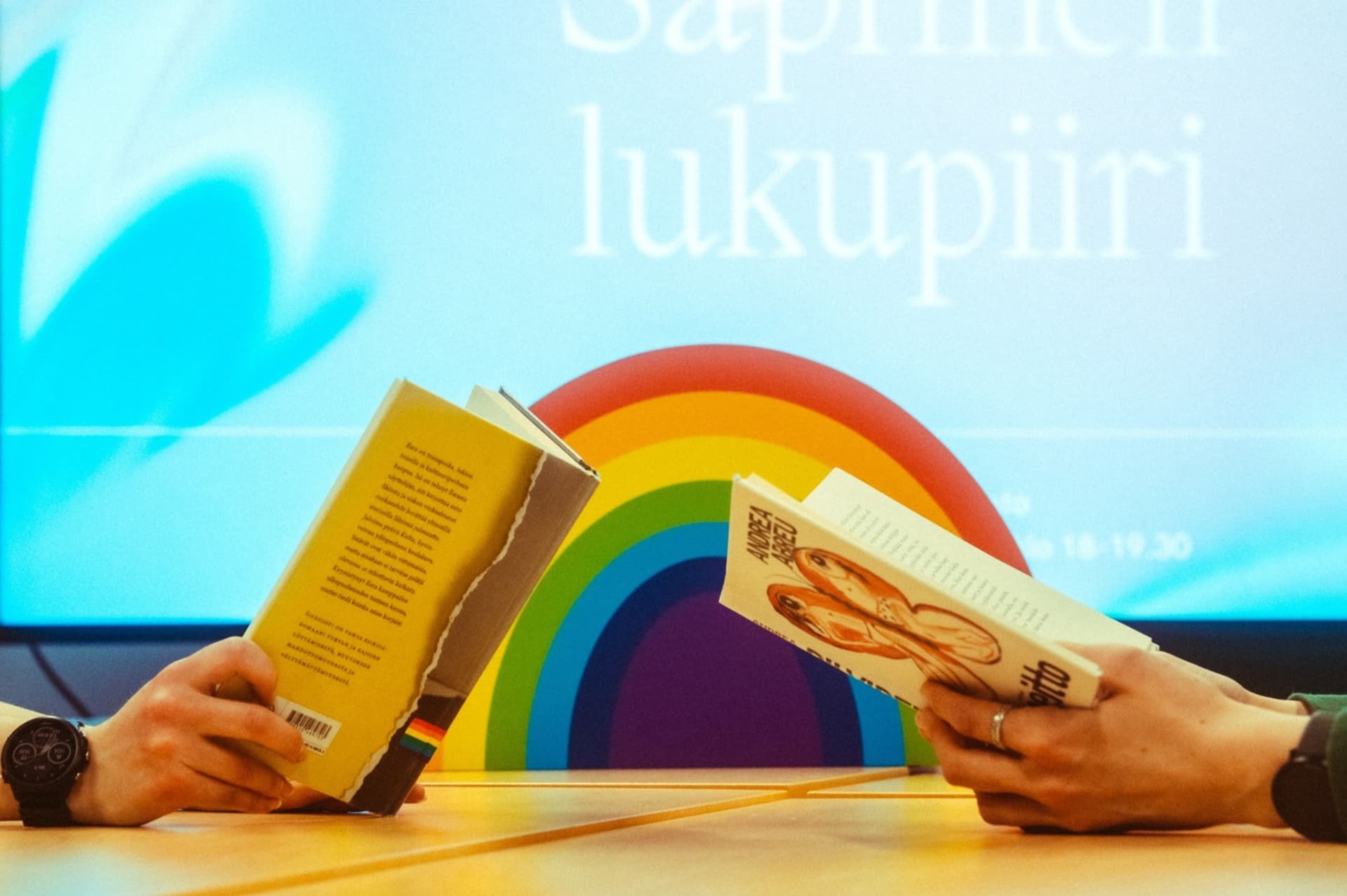 Kahden henkilön kädet, joissa on luettavat kirjat auki. Taustalla sateenkaari sekä näyttö jossa lukee Sapfinen lukupiiri