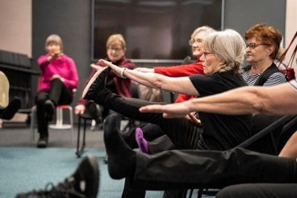 Seniori-ikäiset henkilöt istuvat tuoleilla puoliympyrässä ja ojentavat käsiään ja jalkojaan.