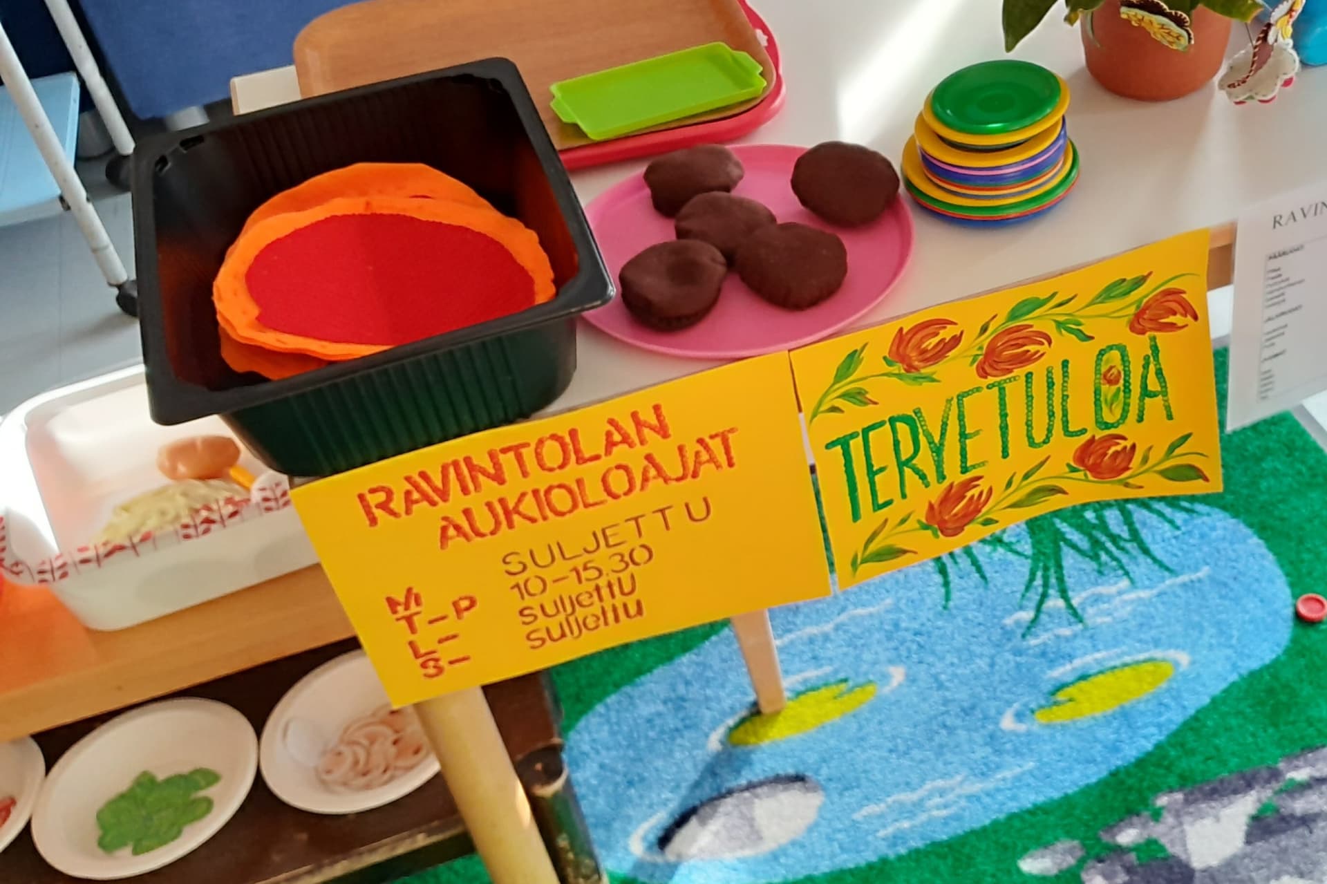 leikkiravintolan aukioloajat ja tervetuloa kyltti sekä leikkiastioita ja -ruokia pöydällä ja hyllyssä, alla näkyy värikäs matto, jossa lampi