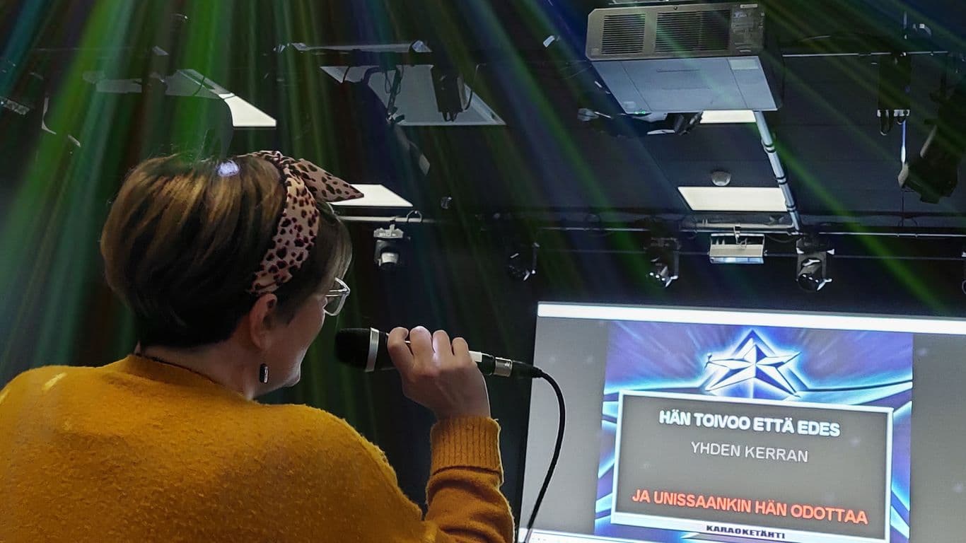Nainen laulaa mikkiin karaokessa ja lukee laulunsanoja näytöltä.