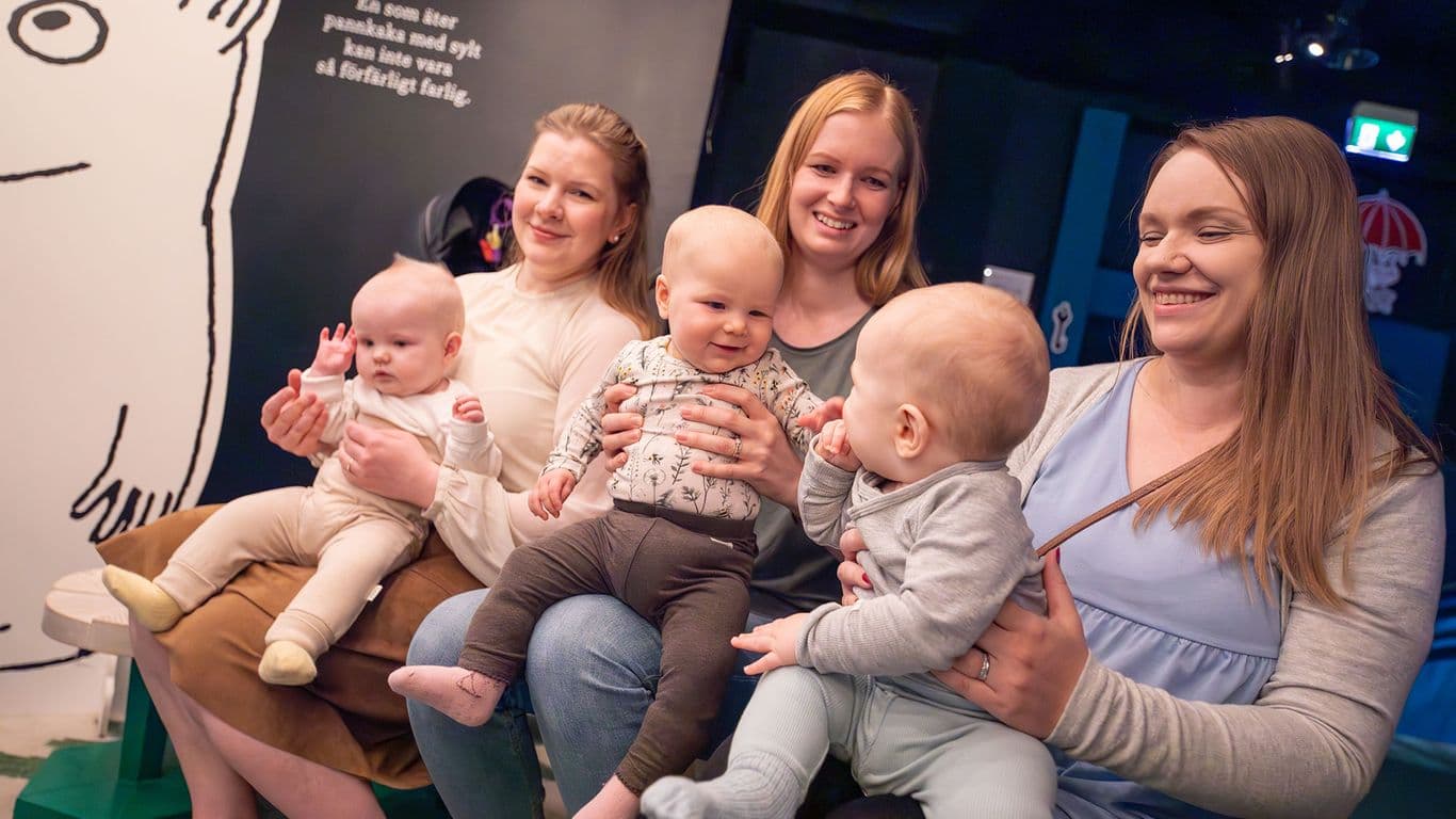 Kolme vauvaa istuu naureskellen äitiensä sylissä.